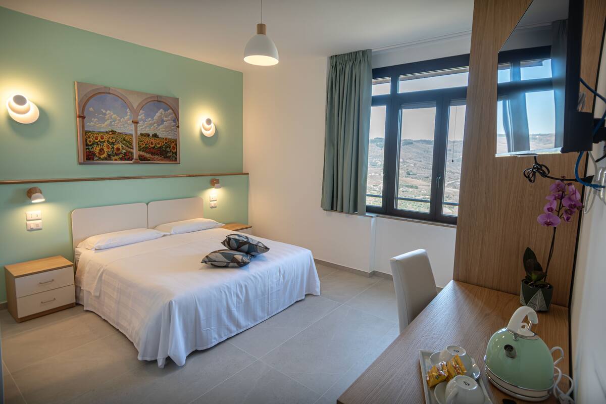A bedroom with a bed, bedside tables, a desk with a chair, a window with a view, and a painting on the wall.