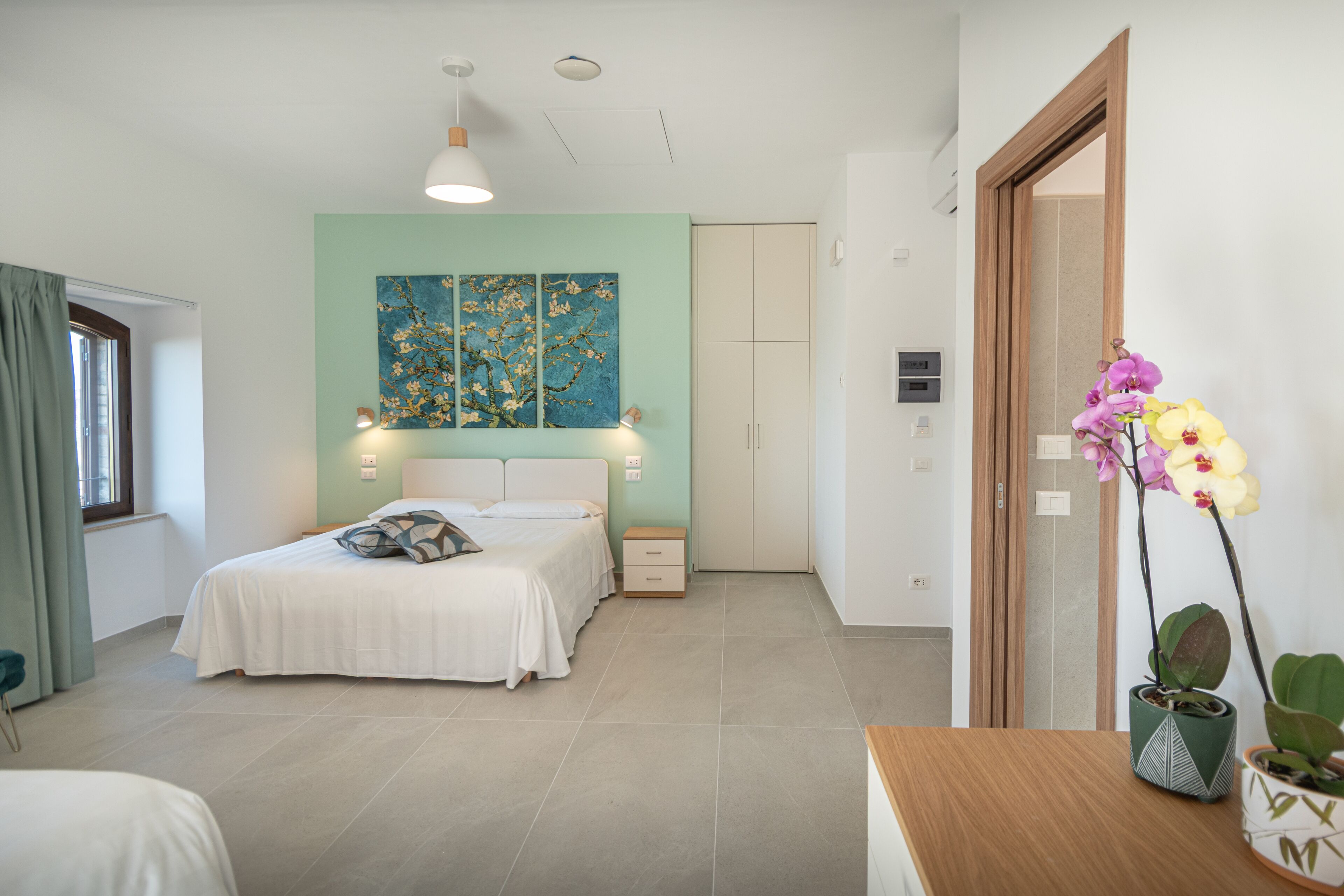 Une chambre à coucher comprenant un lit, des tables de chevet, une commode, une fenêtre avec des rideaux, un luminaire fixé au mur et un miroir.