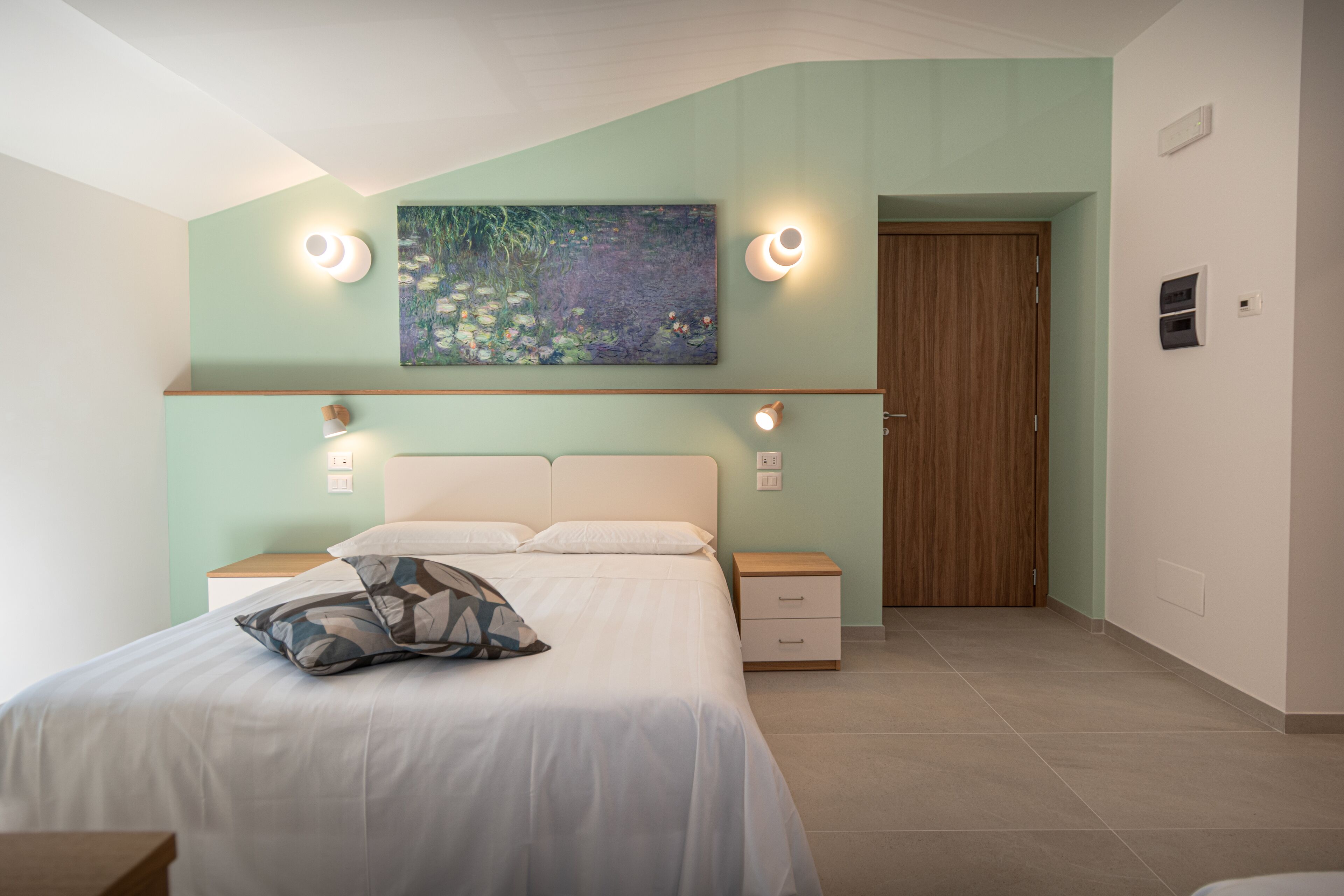 A bedroom with a bed, bedside tables, a nightstand, a wooden door, and a wall-mounted painting.