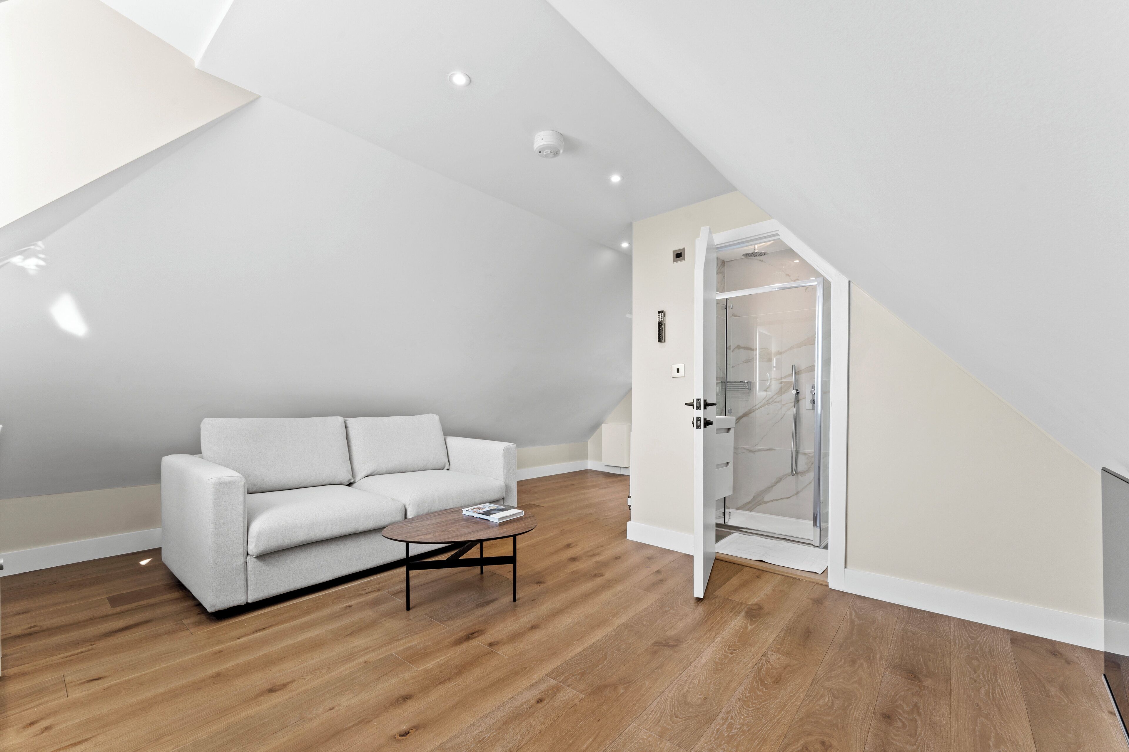 A modern loft bedroom with a sloped ceiling, a white sofa, a wooden coffee table, and a glass shower enclosure.