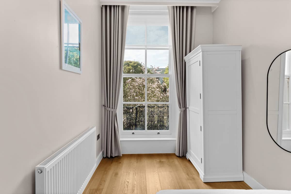 A room with a large window, white radiator, and a white wardrobe.