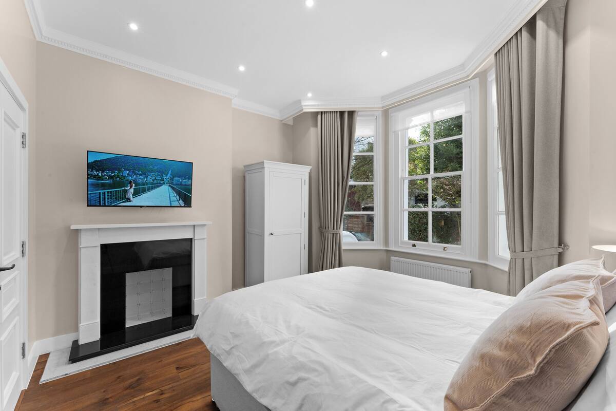 A bedroom with a large bed, a fireplace, a television mounted above it, and a window with curtains.
