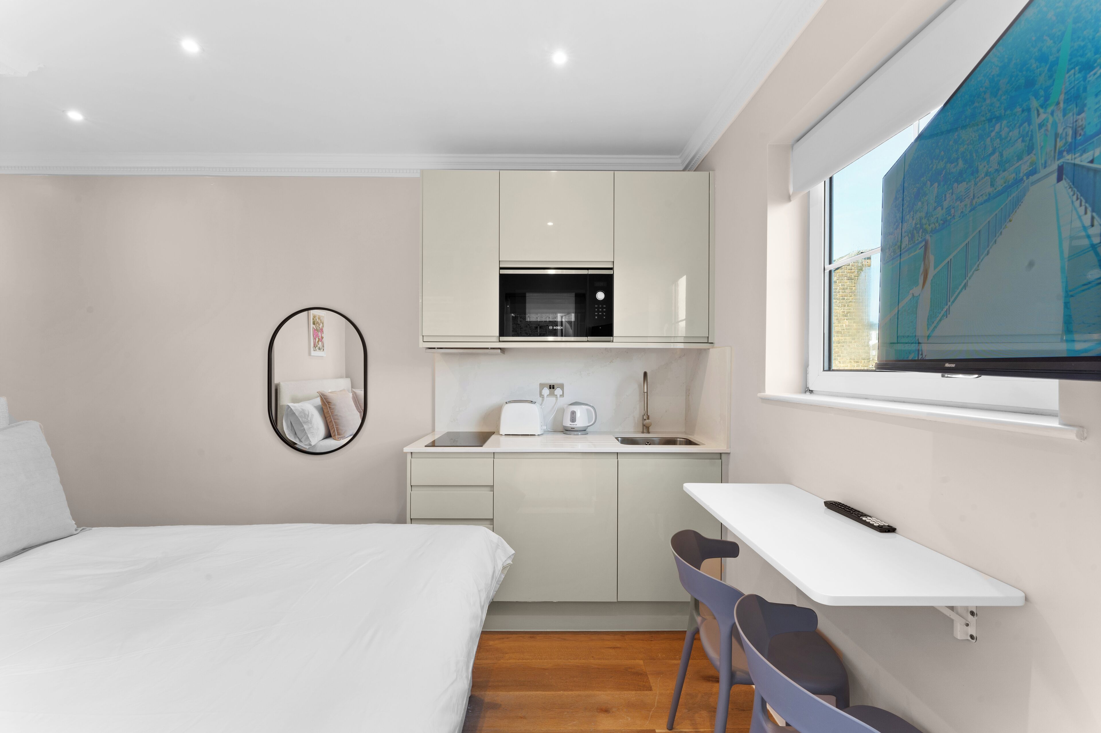 Ein kompaktes Hotelzimmer mit Bett, einer kleinen Küchenzeile, einem Fenster mit Blick auf die Stadt und einem Schreibtisch mit Stuhl.