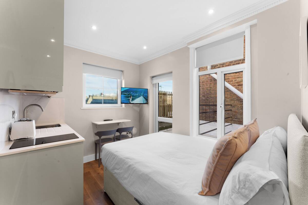 A modern bedroom with a bed, a desk, a TV, and a window with a view of a brick wall and a balcony.