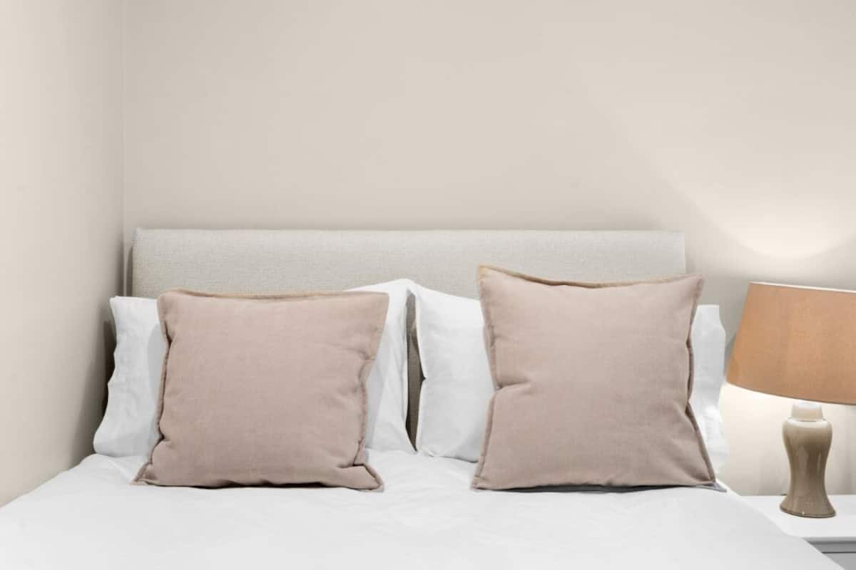 Una cama con almohadas blancas y beige, un cabecero y una lámpara de noche.