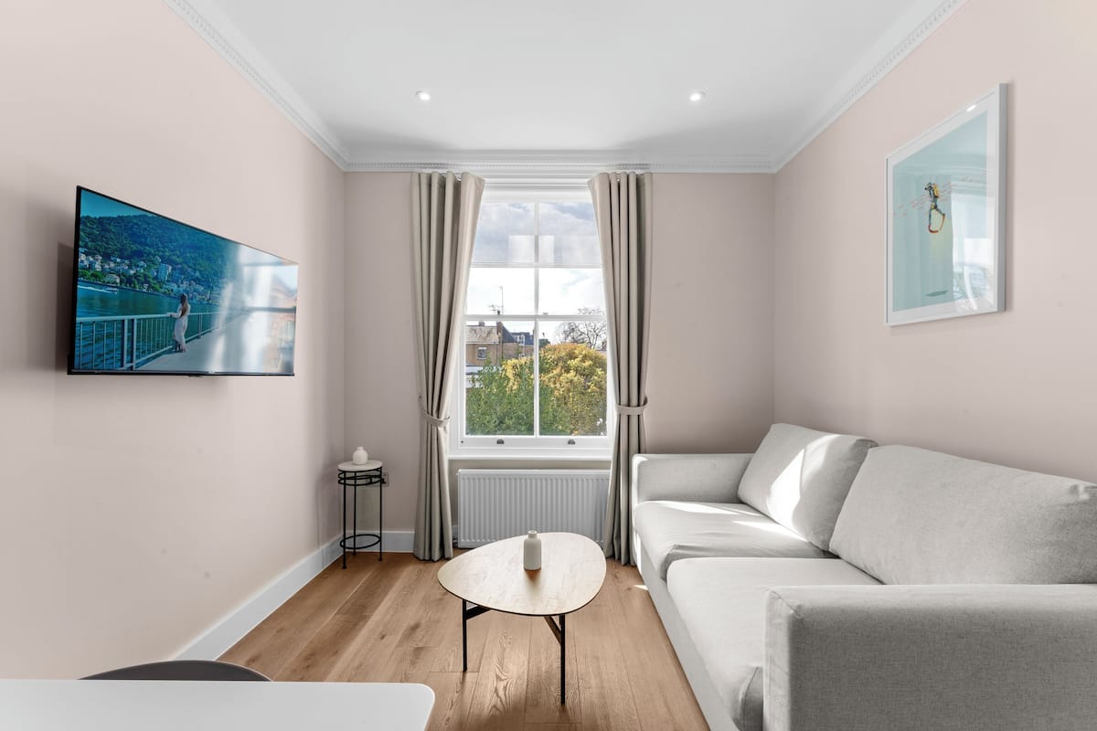 A modern living room with a large window, a sofa, a coffee table, and a TV mounted on the wall.