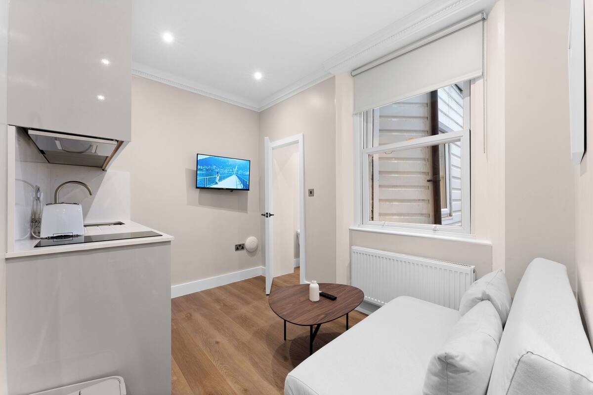 A modern hotel room with a kitchenette, a TV mounted on the wall, a wooden floor, and a white sofa.