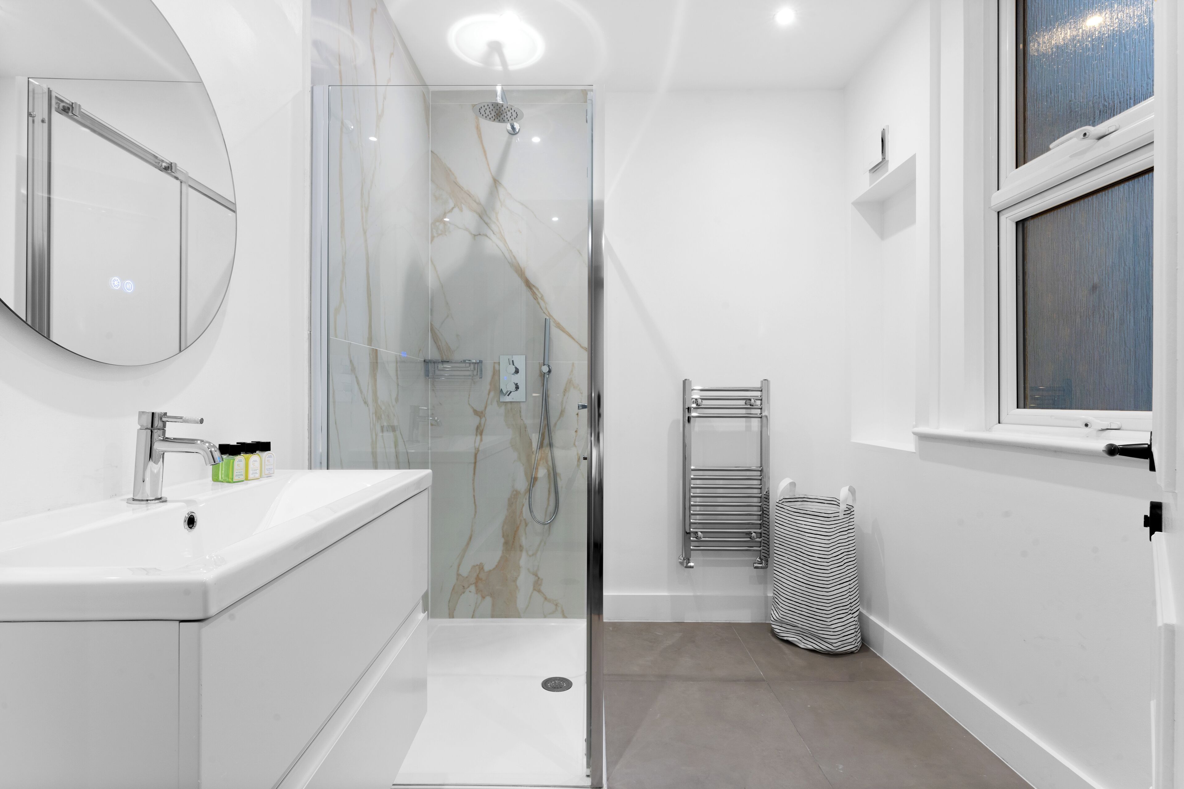 A modern bathroom with a glass-enclosed shower, a white sink with a mirror, and a window.