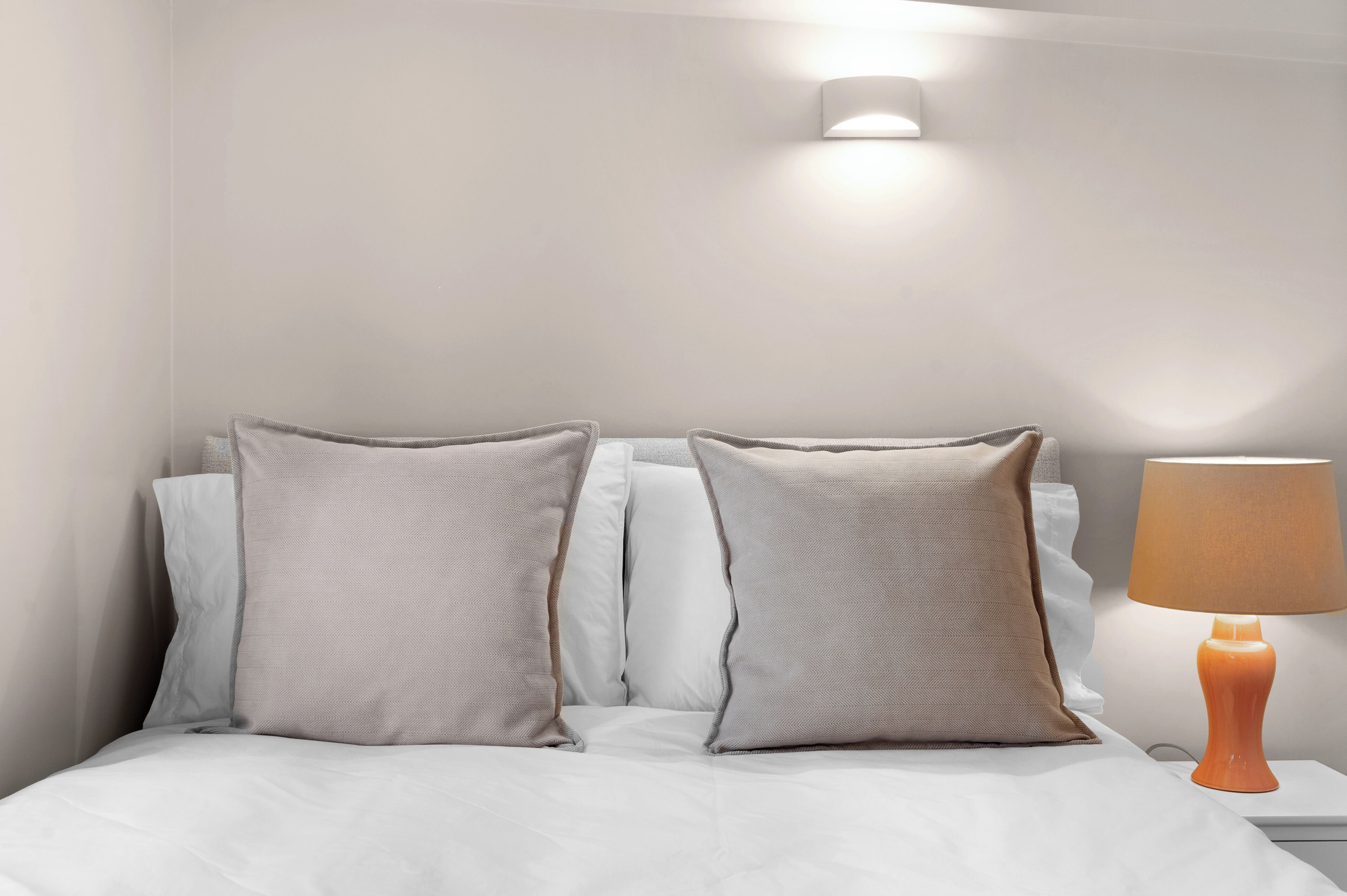 Een net opgemaakt bed met witte lakens en twee kussens, een nachtkastje met een lamp en een wandlamp.