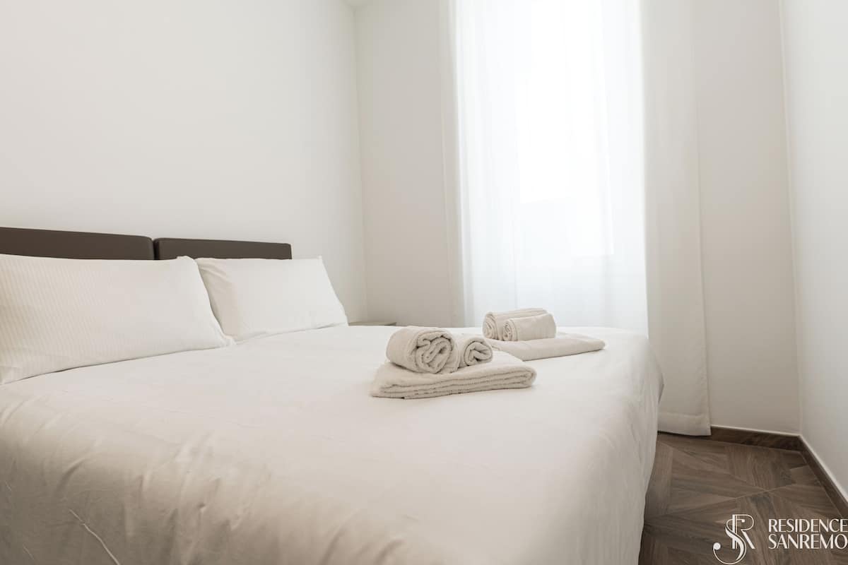Un lit bien fait, avec du linge de lit blanc et des serviettes pliées, une tête de lit en bois et une fenêtre ornée de rideaux légers.