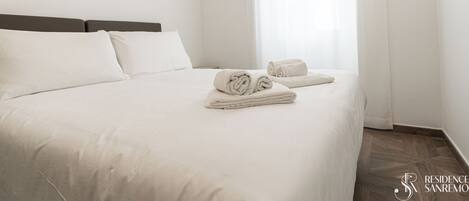 A neatly made bed with white linens and rolled towels, a wooden headboard, and a window with sheer curtains.