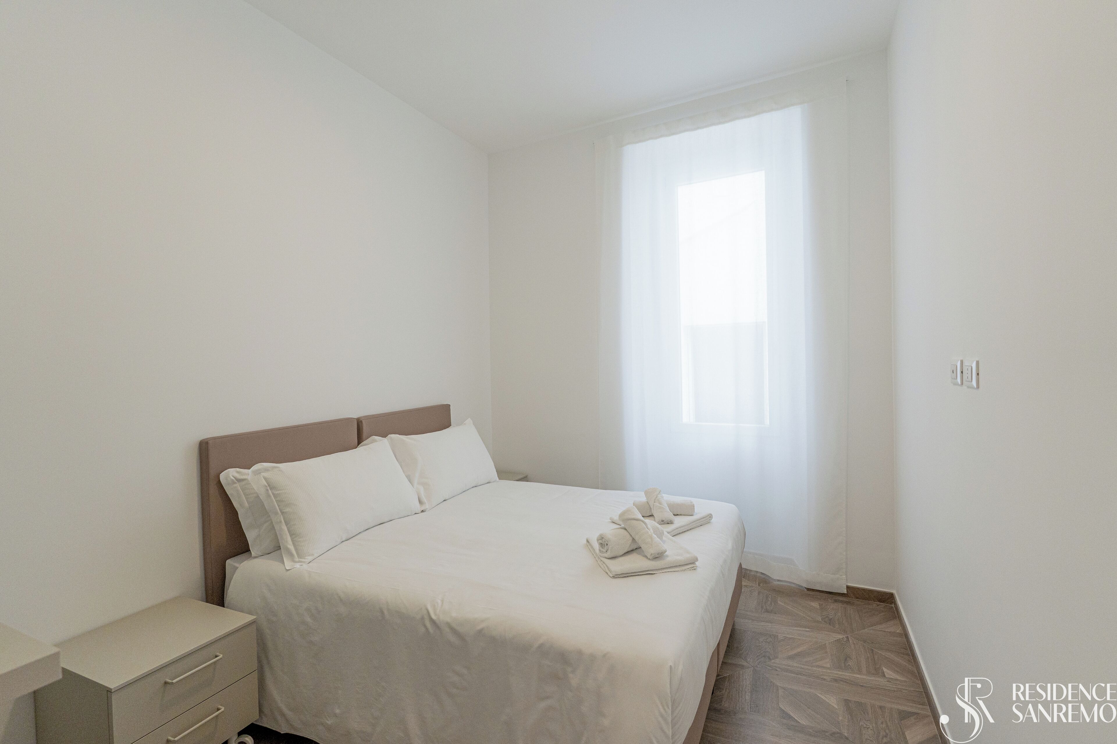 Ein schlicht eingerichtetes Schlafzimmer mit Bett, Nachttisch und Fenster.