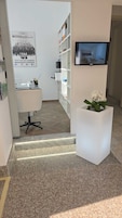 Un espacio de oficina moderno con un escritorio blanco, una silla y una estantería. Un televisor está montado en la pared y una planta en maceta en el suelo.