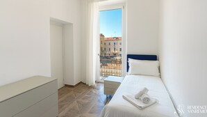 Et moderne hotelværelse med seng, lille natbord, garderobeskab og en balkon med udsigt til bygninger.