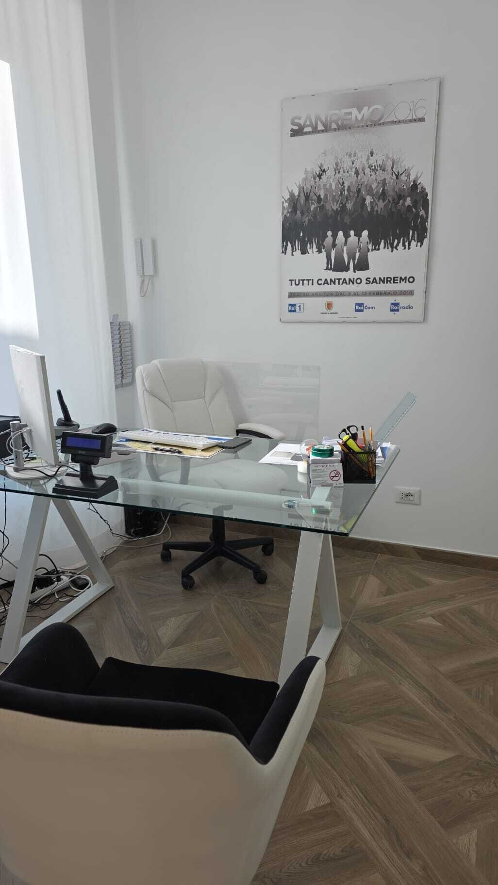A modern office with a glass desk, a computer, and a chair. There's a poster on the wall and a telephone on the wall.