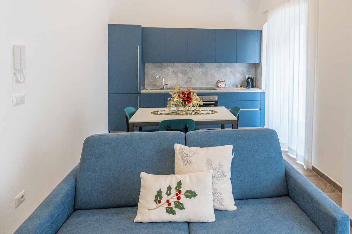 Un salon moderne avec un canapé bleu, deux coussins décoratifs, une table à manger et un coin cuisine doté de meubles bleus et d’un plan de travail blanc.