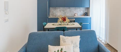 A modern living room with a blue sofa, two decorative pillows, a dining table, and a kitchen area with blue cabinets and a white countertop.