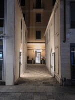 Un pasillo con una puerta central que da a una entrada iluminada, flanqueado por dos edificios con ventanas y una cámara de seguridad.