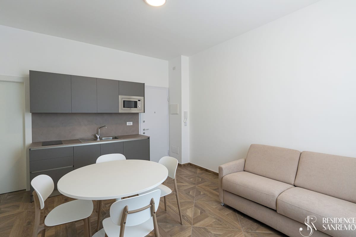Une cuisine moderne avec des meubles gris, un four à micro-ondes et un évier. On y trouve une table ronde avec des chaises et un canapé beige dans l’espace salon.