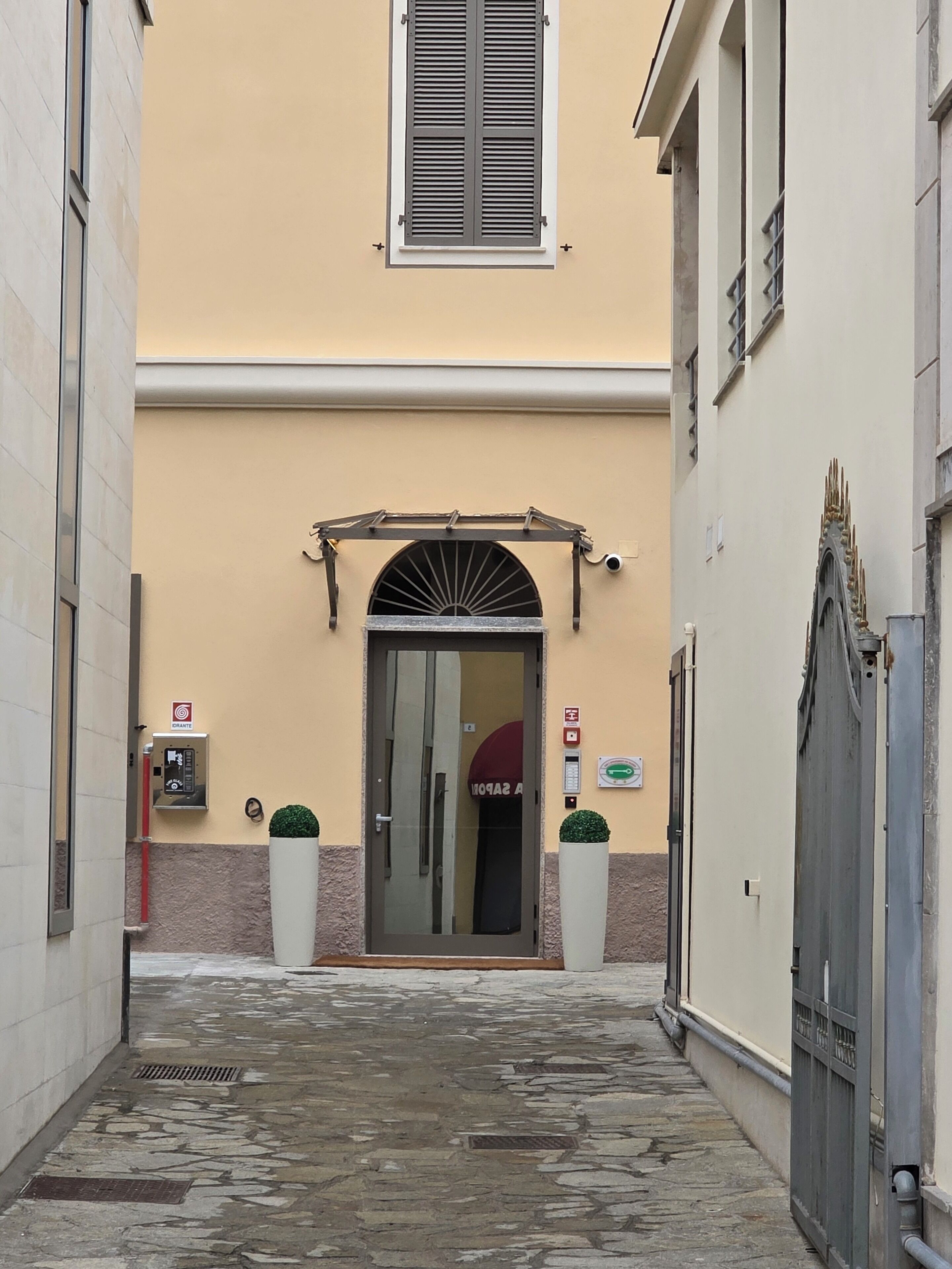 Ein schmaler Gang führt zu einem Gebäude mit Glastür und einem kleinen Rundbogenfenster darüber. Vor der Tür stehen je ein Blumentopf auf beiden Seiten, und links an der Wand ist ein Brandmelder angebracht.