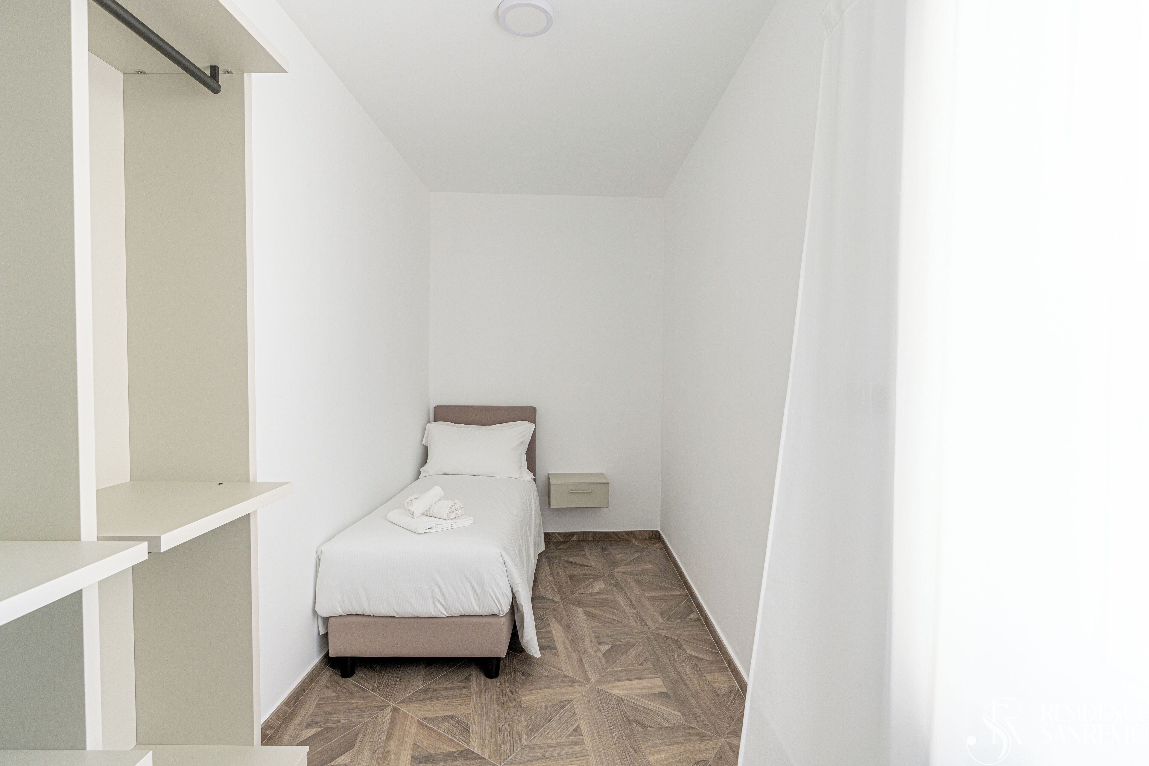 Une chambre étroite aux murs blancs, avec un lit, une table de chevet et une étagère intégrée.