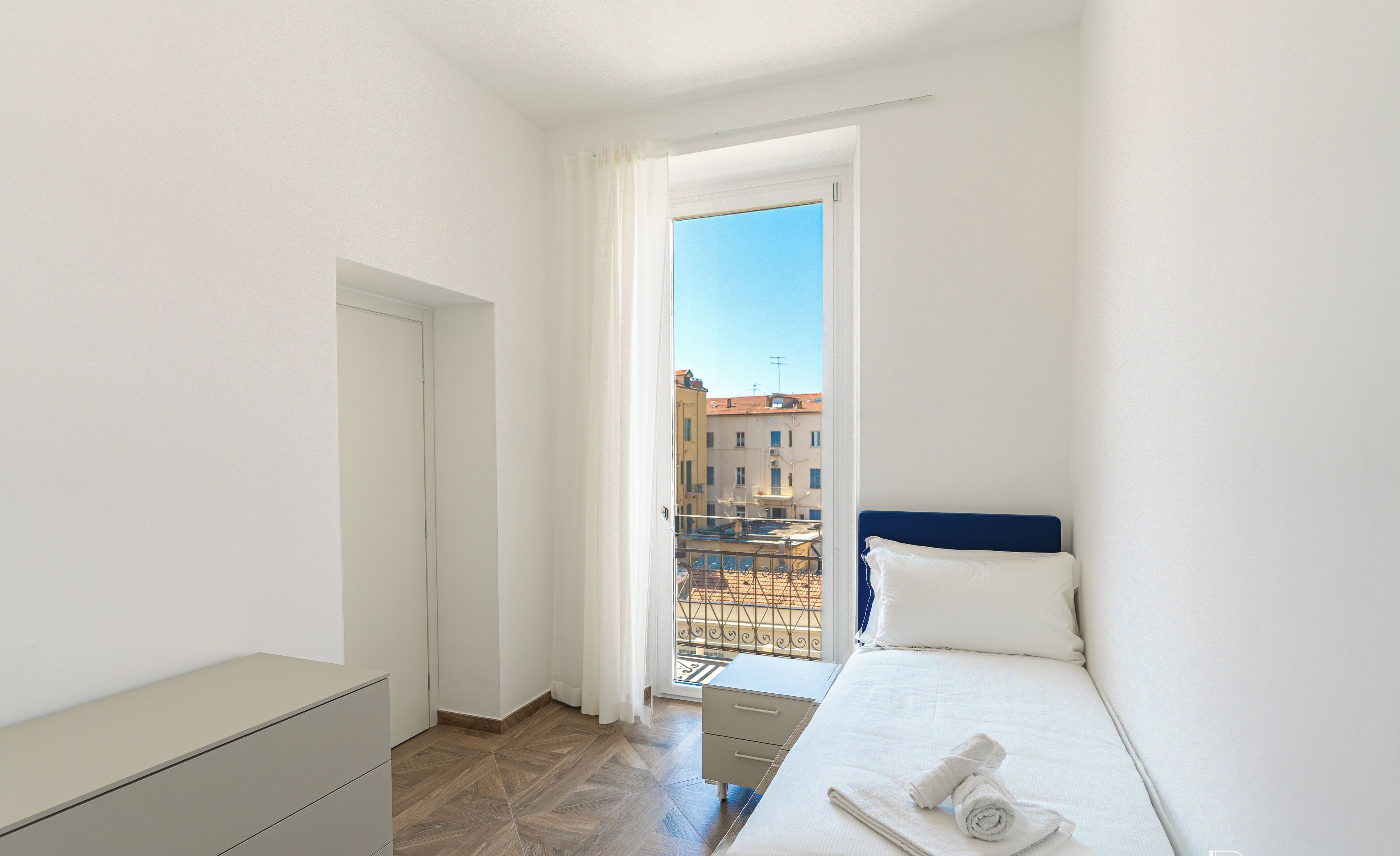 A modern hotel room with a bed, a small bedside table, a wardrobe, and a balcony with a view of buildings.