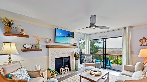 A spacious living room with a fireplace, comfortable seating, a television, and a balcony with a view of the beach. - Marsh 2 the Beach, 2 Bedroom Island Rental (Saint Simons Island)
