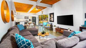A modern living room with a large grey sectional sofa, a flat-screen TV, and a kitchen in the background. - 30+ Stay! W/Pool, Hot Tub, Large Patio, Ping Pong (Palm Springs)