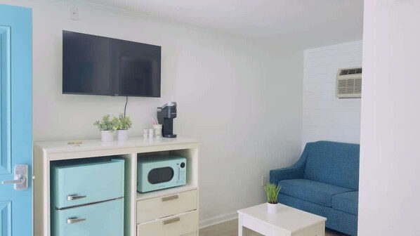A room with a flat-screen TV, a blue sofa, a small white table, a kitchenette with a fridge and microwave, and a door with a handle.