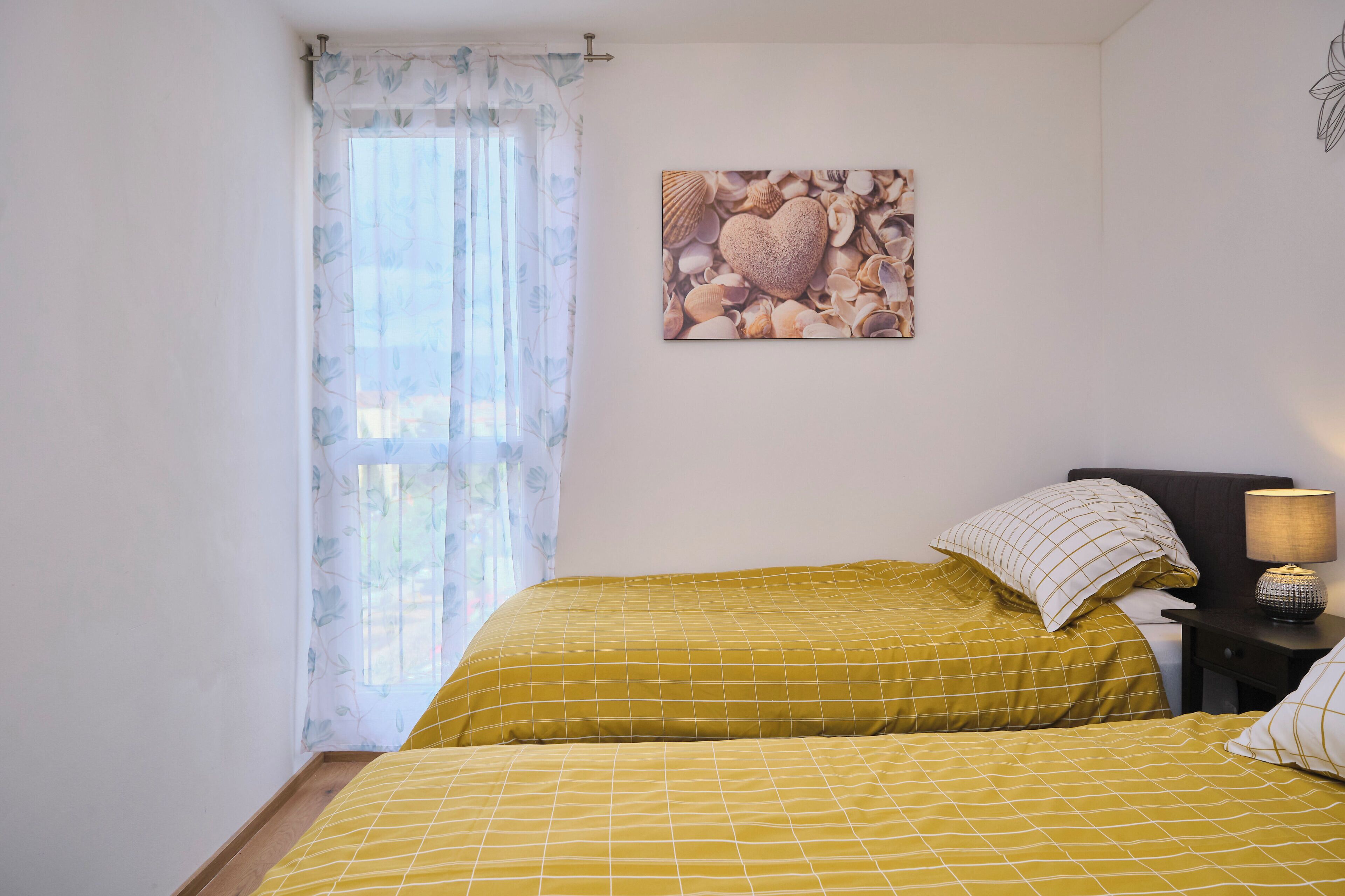 A room with two beds, a nightstand with a lamp, a window with floral curtains, and a wall art of seashells.