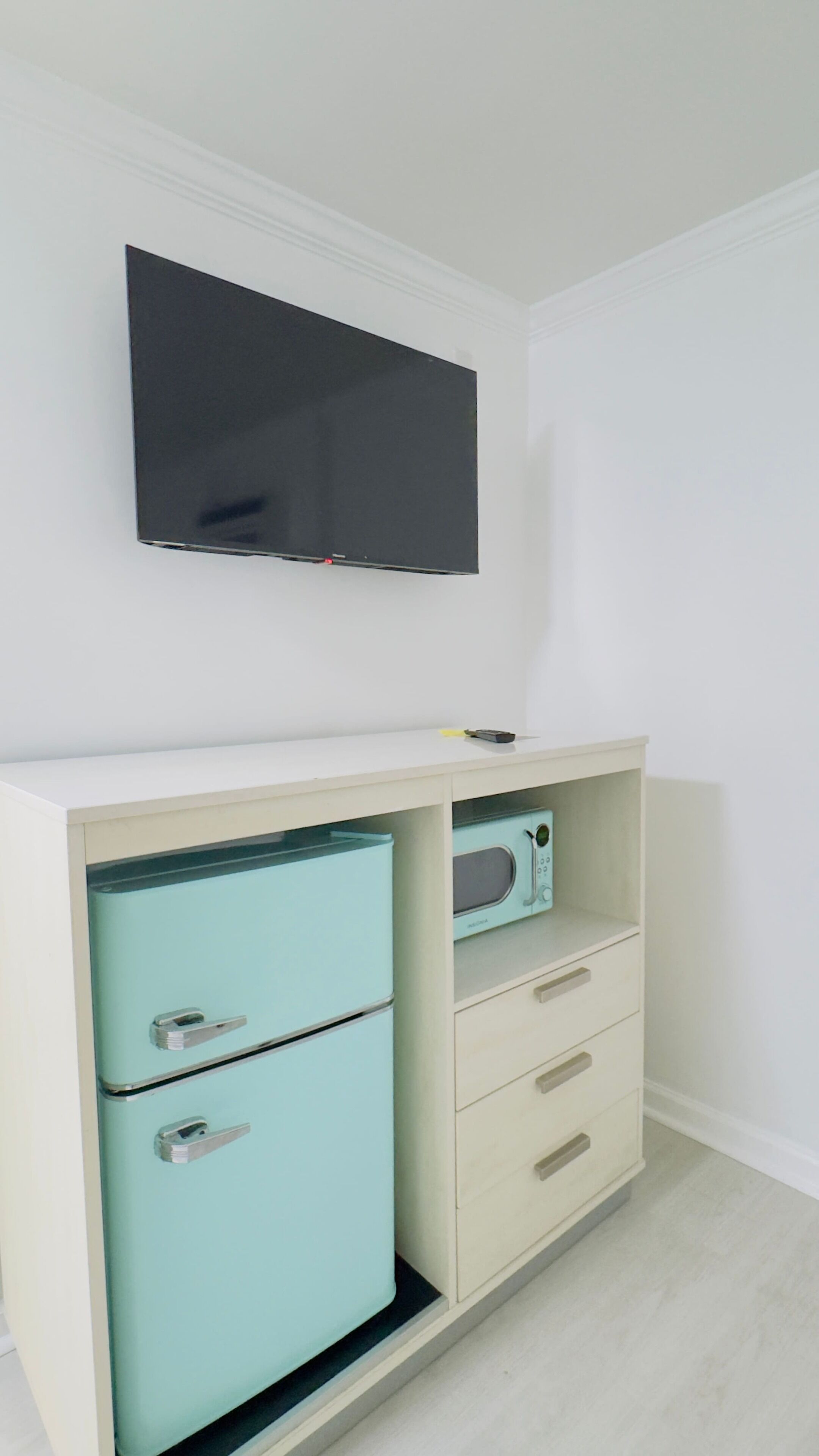 A room with a white wall, a flat-screen TV mounted above a light blue cabinet with a microwave, and a small drawer unit.