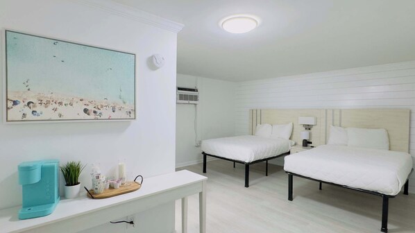 Habitación de hotel con dos camas, una mesita consola blanca con cafetera, un cuadro de una playa y una lámpara en el techo.