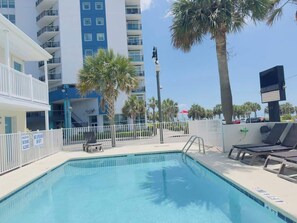 A swimming pool with lounge chairs, a Starbucks sign, and a tall building in the background. - Boutique on the beach, Studio #120 + Pool (Myrtle Beach)