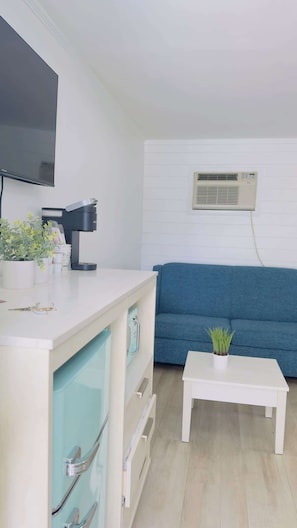 Una sala de estar moderna con televisor de pantalla plana, cafetera, sofá azul, mesa de centro blanca y equipo de aire acondicionado.