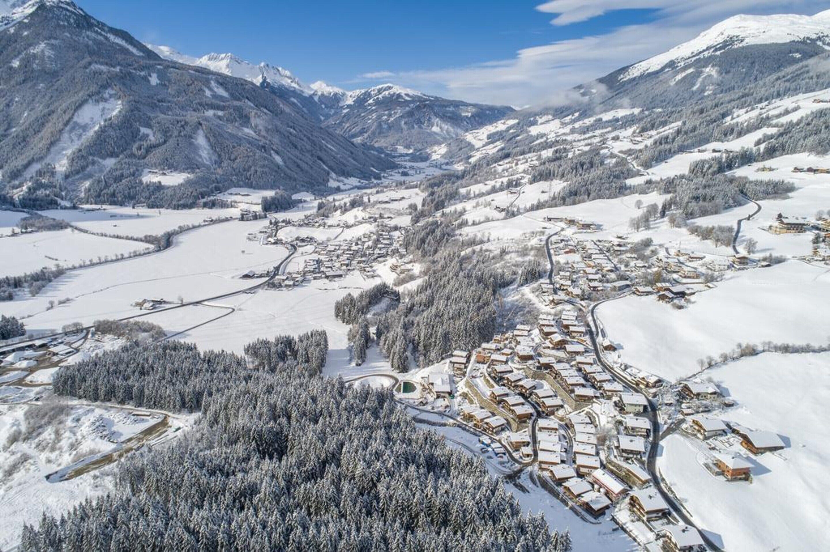 Un pueblo cubierto de nieve con edificios dispuestos en un patrón circular alrededor de una zona verde central, rodeado de montañas boscosas.