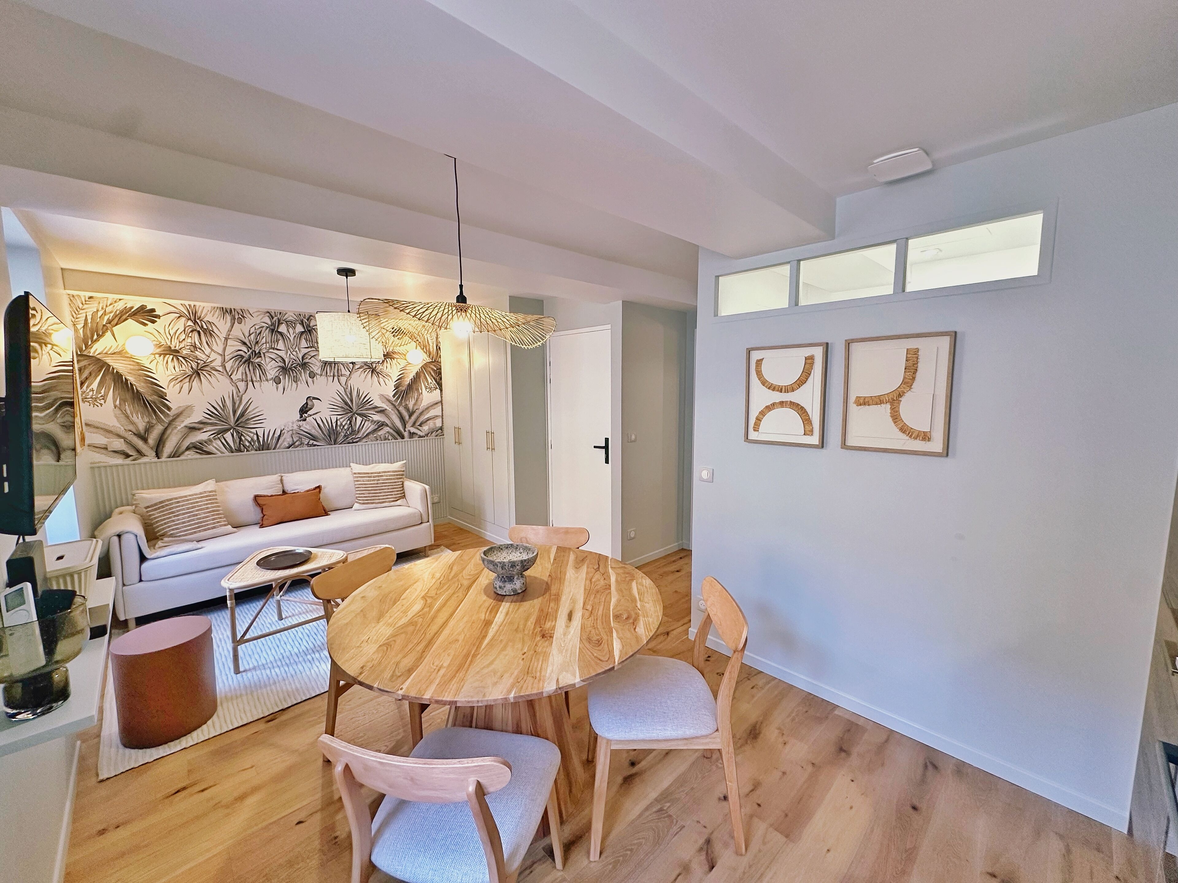 Un salon moderne avec une table de salle à manger ronde en bois, un canapé et un mur recouvert d’un papier peint aux motifs tropicaux.