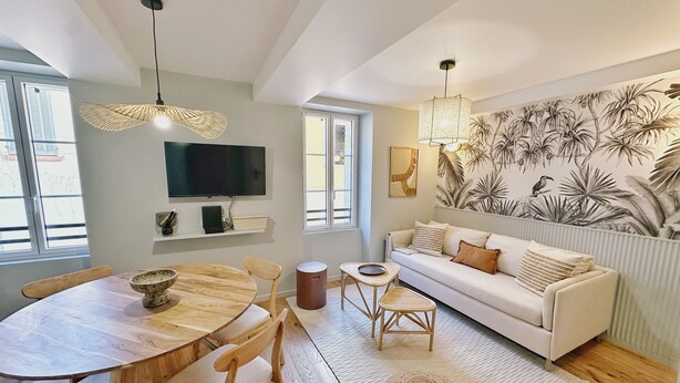 Ein modernes Wohnzimmer mit einem runden Holztisch, zwei Holzstühlen, einem weißen Sofa mit Kissen, einem gewebten Pouf und einem an der Wand montierten Fernseher.