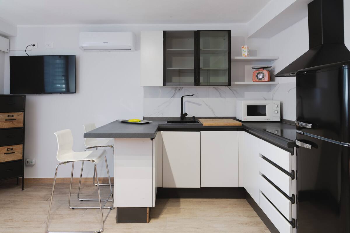 Una cocina moderna con armarios blancos, una encimera negra, un microondas, una estufa y un televisor instalado en la pared.