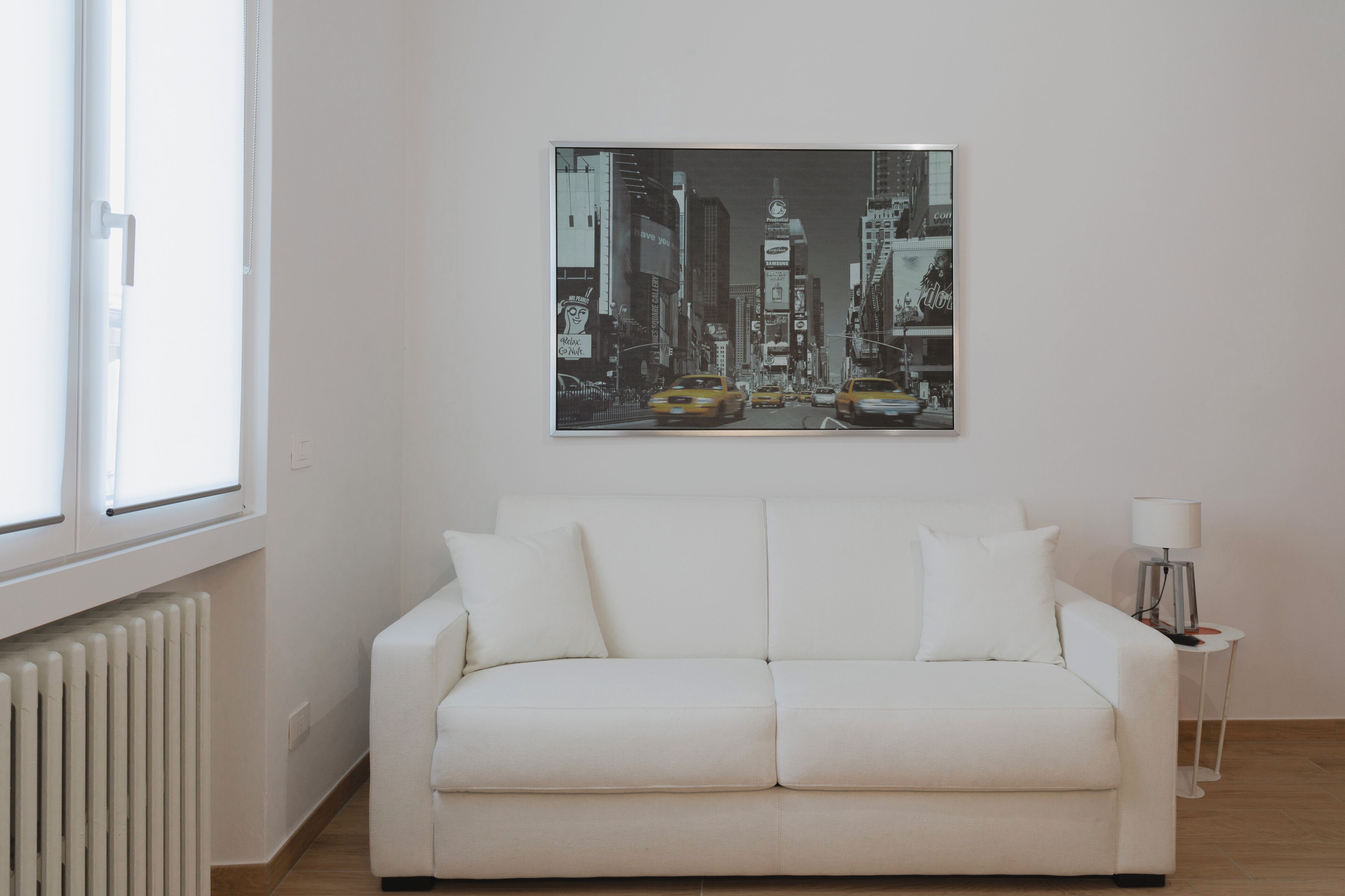 A white sofa in a room with a large cityscape painting on the wall, a window, and a small table with a lamp.