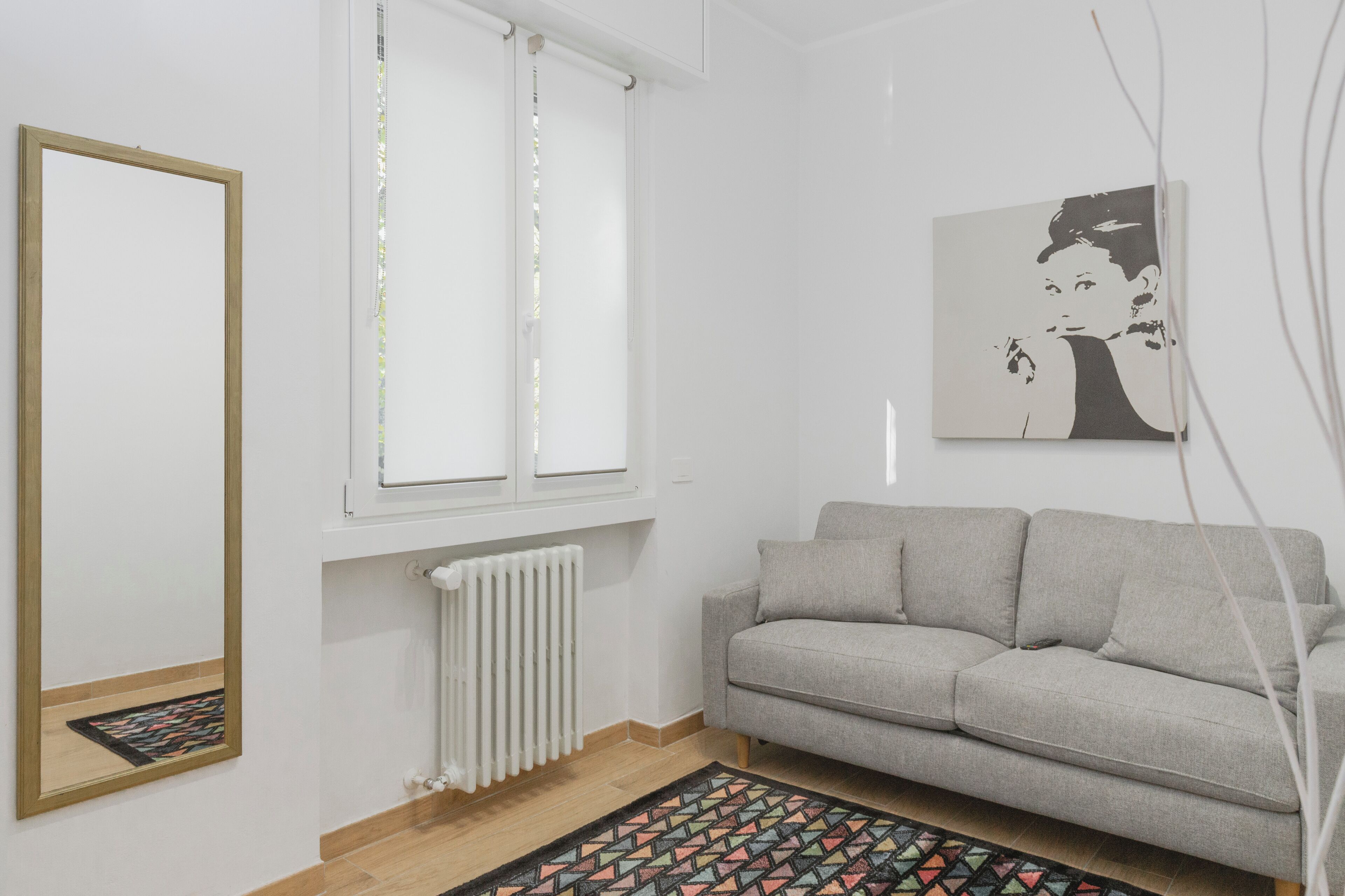 En stue med en grå sofa, en hvid radiator, et vindue med skodder, et spejl og et billede i ramme på væggen.