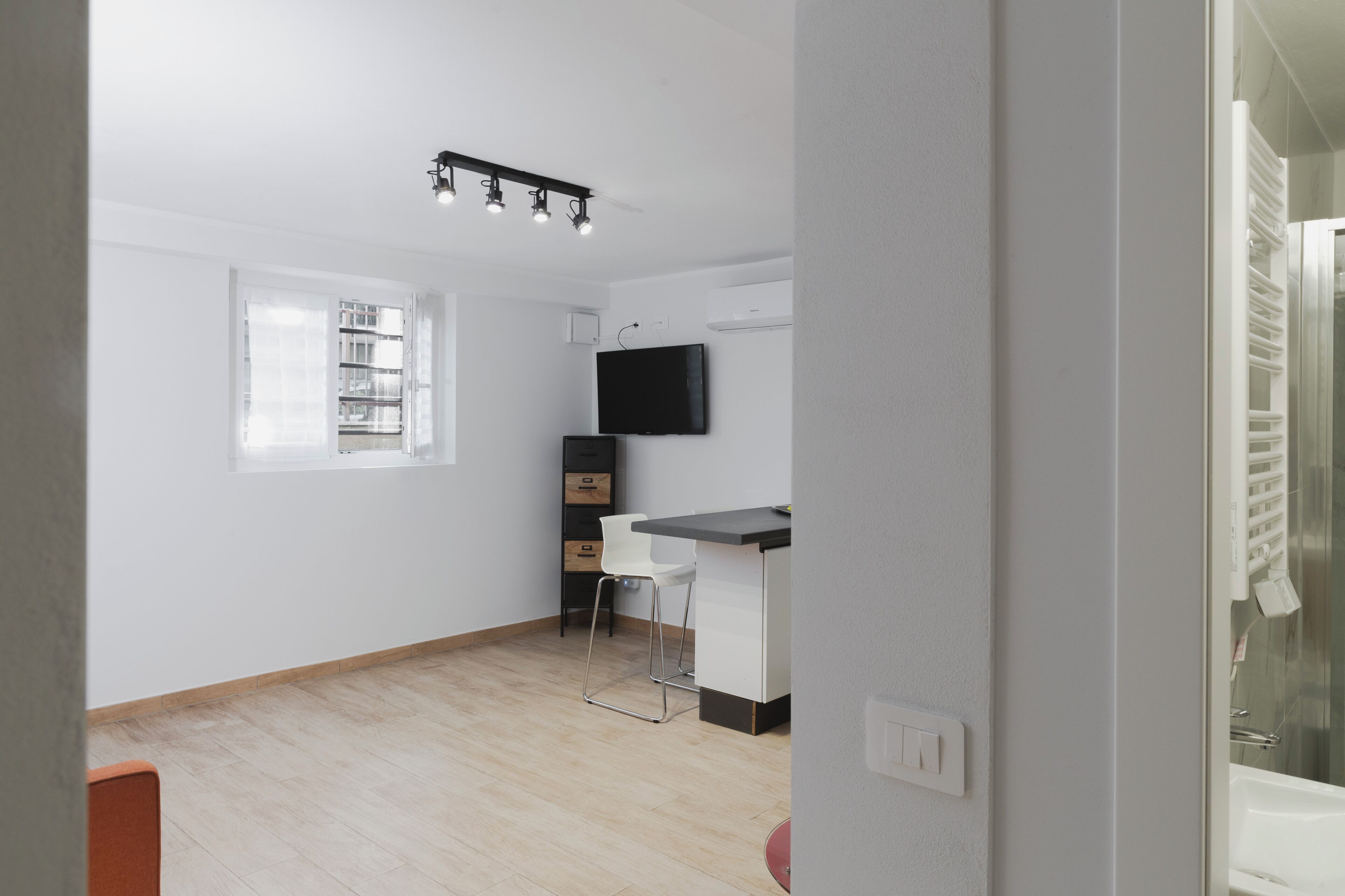 Eine moderne Küche mit schwarzem Arbeitsplatz, weißen Schränken und einem eingebauten Ofen. Ein Fenster mit Jalousien, ein weißer Stuhl und ein an der Wand befestigtes Fernsehgerät sind ebenfalls vorhanden.