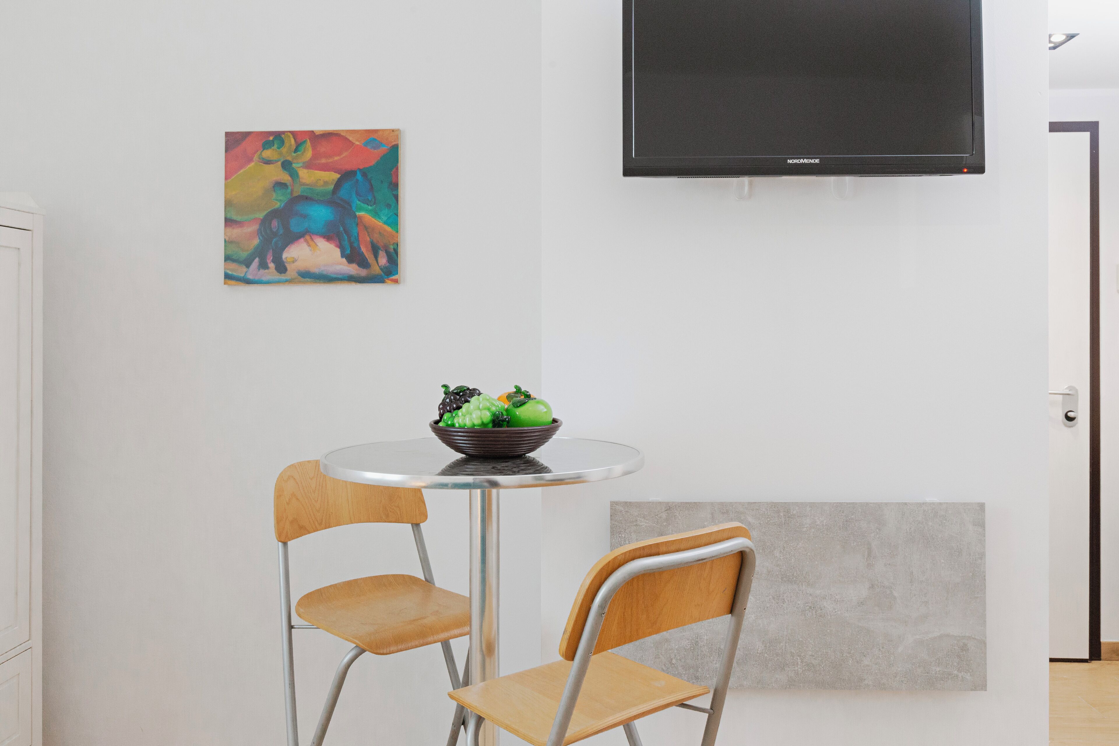 A modern room with a round table, two wooden chairs, a bowl of fruit, a colorful abstract painting, and a flat-screen TV mounted on the wall.