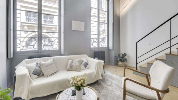 A modern living room with a white sofa, a round coffee table, and a chair. There is a staircase to the right and large windows with a view of buildings. - Sun-filled Loft in the heart of the Marais (Paris)