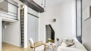A modern living room with a white sofa, a wooden chair, and a coffee table. There is a ladder leading to a lofted area and a staircase to another level. - Sun-filled Loft in the heart of the Marais (Paris)