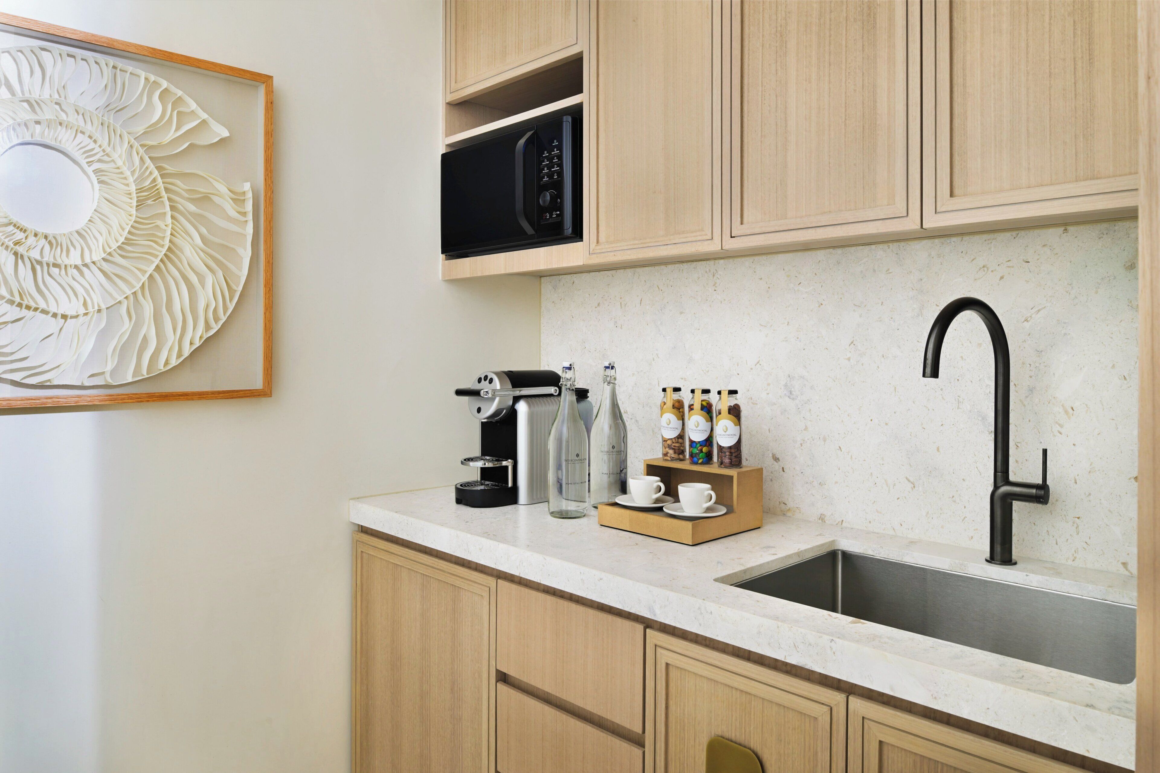Cozinha com armários de madeira, pia de aço inoxidável, cafeteira, micro-ondas e um painel decorativo na parede.