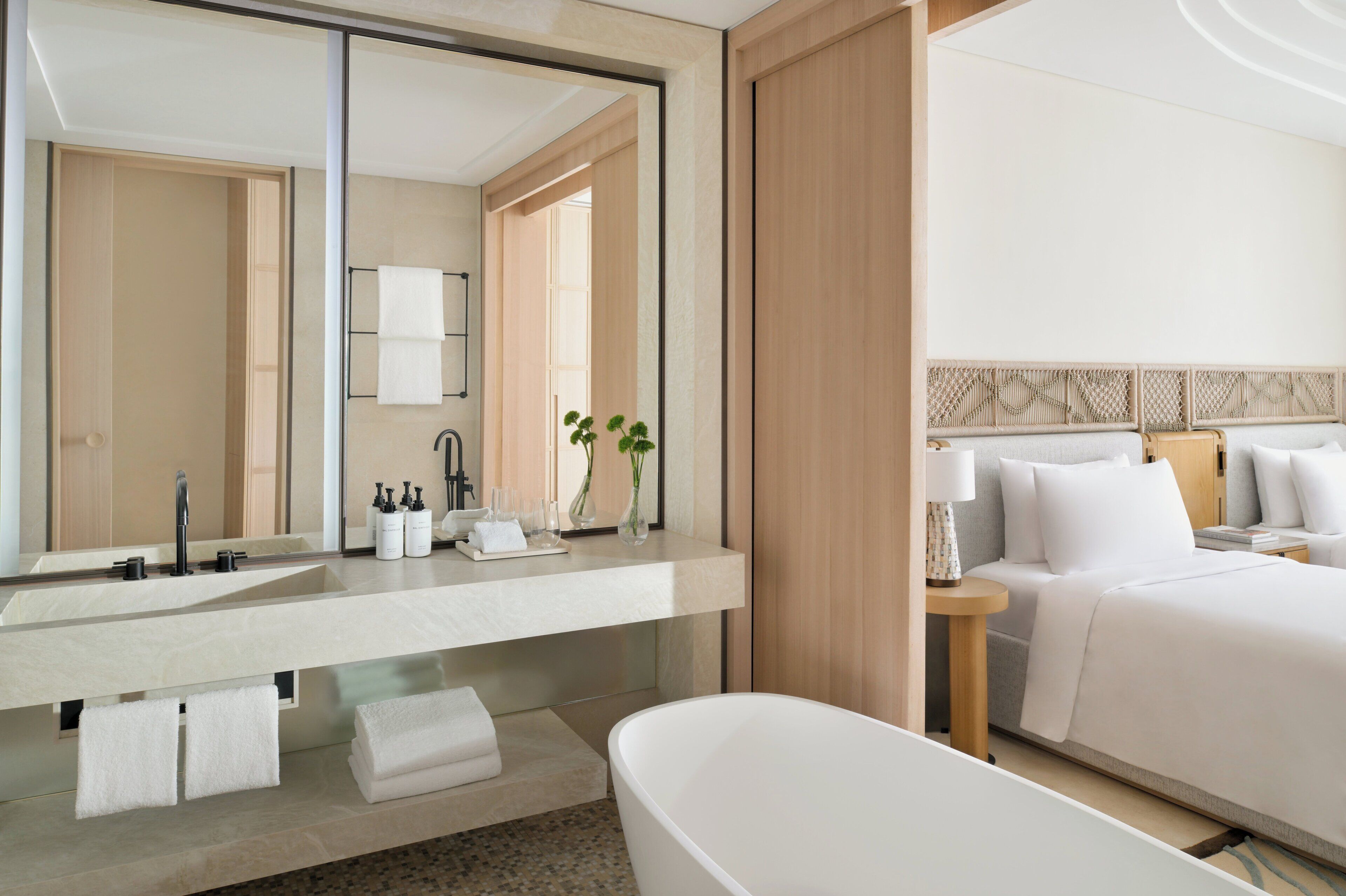 Un baño de hotel moderno con un espejo grande, lavamanos dobles, una bañera y una mesita de noche con lámpara.
