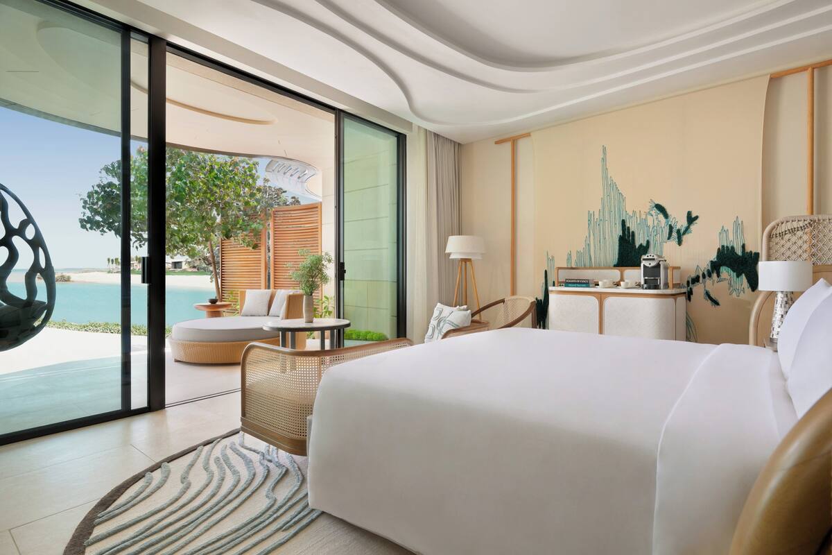 A modern hotel room with a large bed, a dining area, a view of the ocean, and a wall with a cityscape painting.