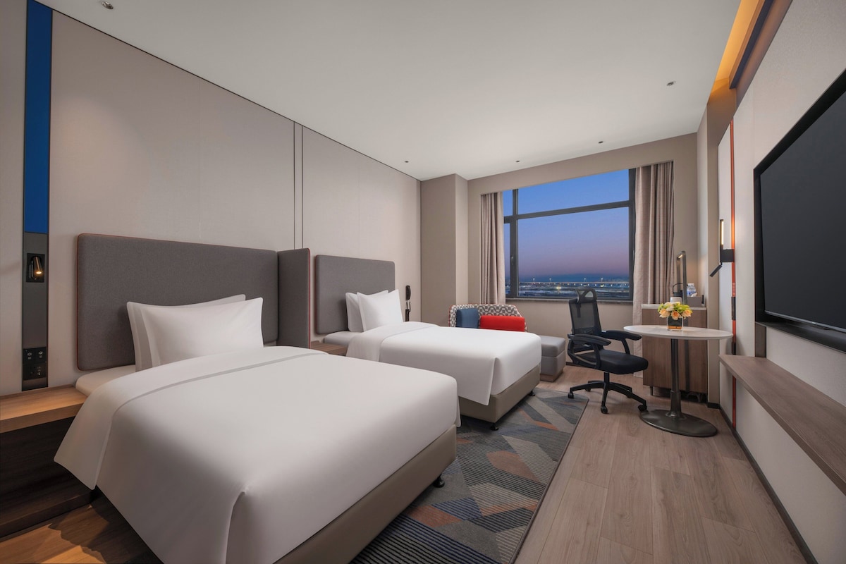 A hotel room with two beds, a desk, a chair, a TV, and a large window with a view.