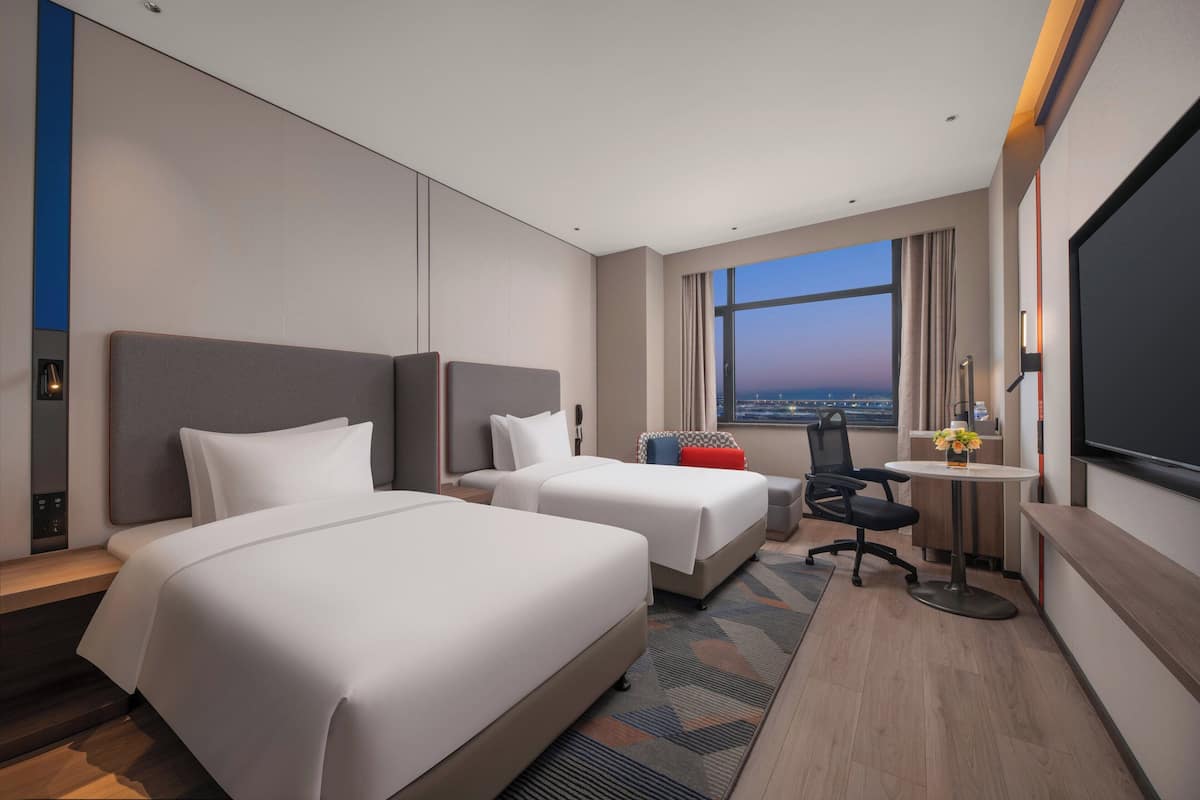 Habitación de hotel con dos camas, un escritorio, una silla, un televisor y un ventanal con vistas.