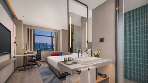 A modern hotel room with a large bed, a desk, a bathroom with a large mirror, and a view of the cityscape.